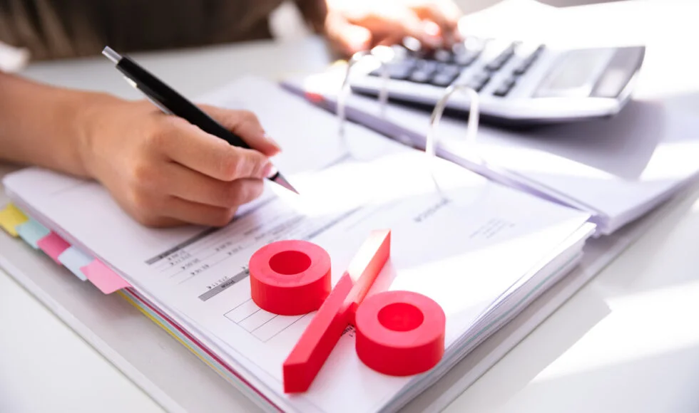 Large red percentage symbol in the foreground with a person performing financial calculations and taking notes in the background.