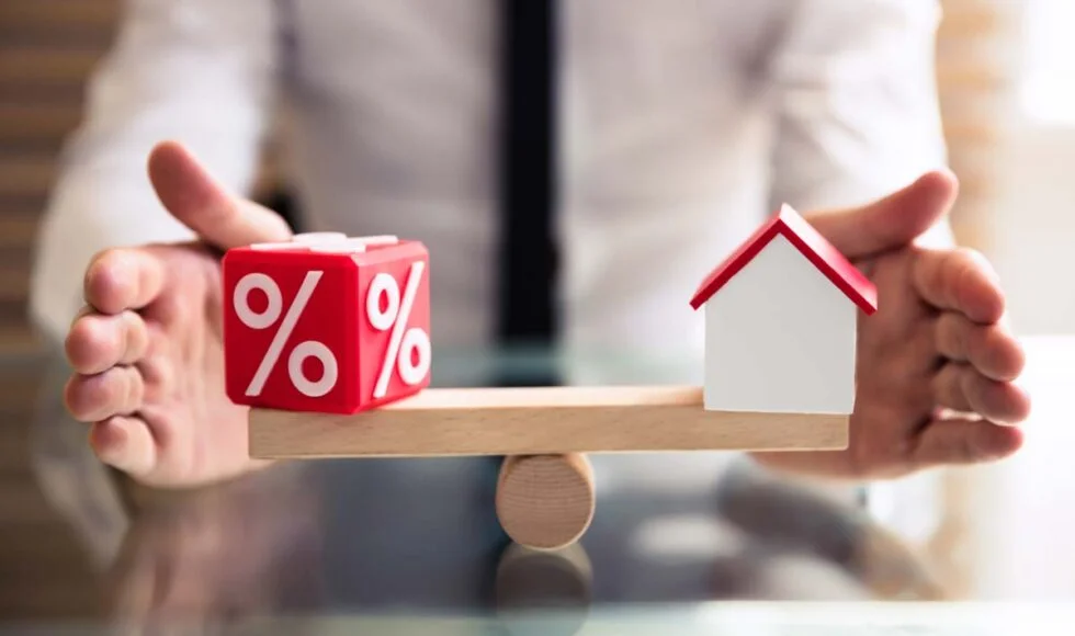 Hands balancing a wooden seesaw with a red percentage block on one side and a miniature white house model on the other, symbolizing the relationship between interest rates and housing.