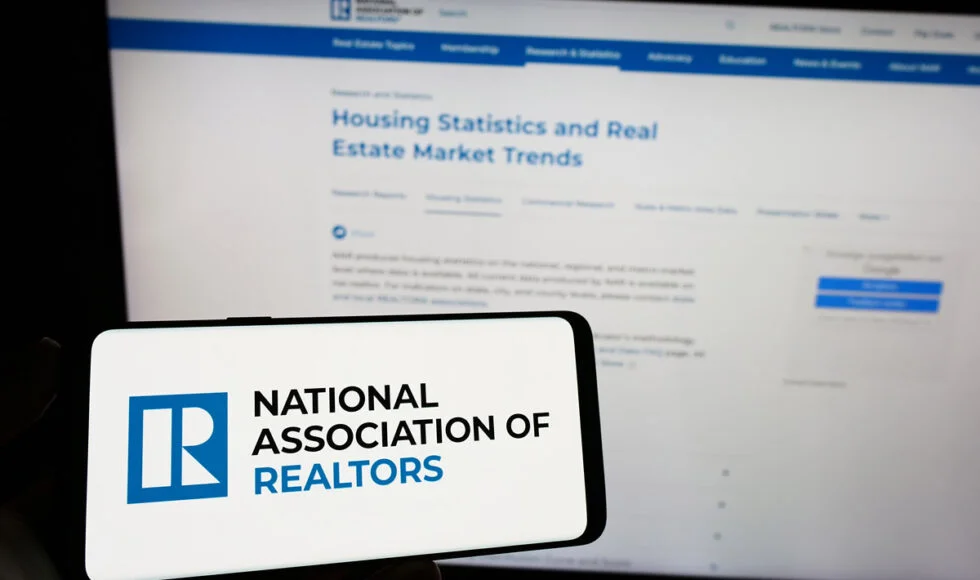 National Association of Realtors (NAR) logo displayed on a smartphone screen with housing statistics visible on a monitor in the background.